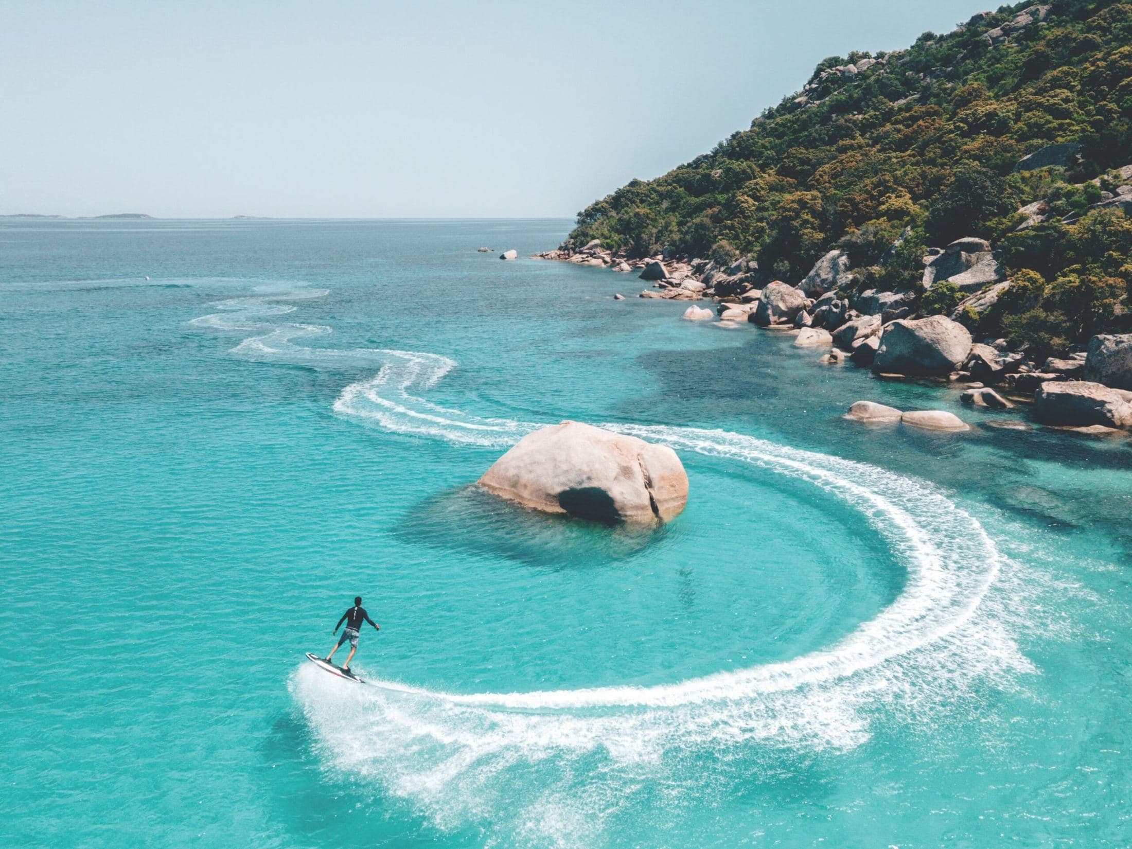 Aerial view of an electric jet board carving a circle around a granite outcrop in turquoise water
